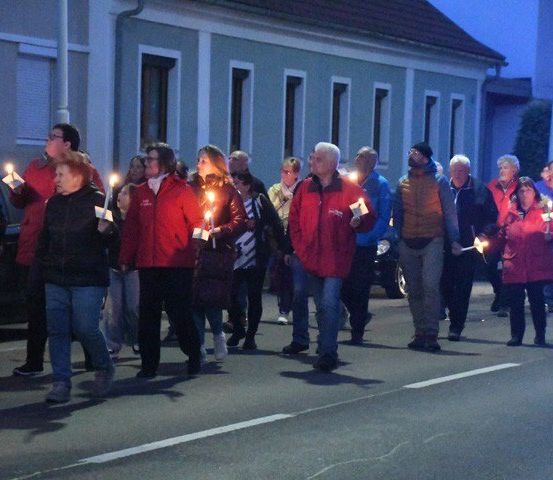 Eine Gruppe von Menschen, alle in roten Jacken gekleidet, gehen bei Nacht die Straße entlang und halten Kerzen in den Händen.