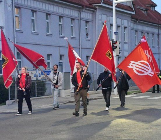 Eine Gruppe von Menschen geht mit roten Fahnen eine Straße entlang, mit einem Gebäude und Ampeln im Hintergrund.