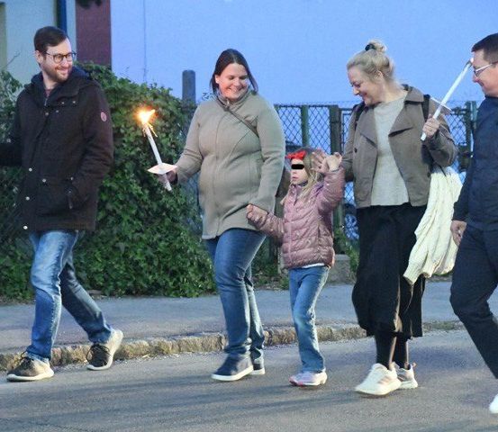 Eine Familie mit fünf Personen geht zusammen, jede hält eine brennende Kerze, auf einer Straße bei Nacht. Sie sind in warme Kleidung gekleidet und lächeln.