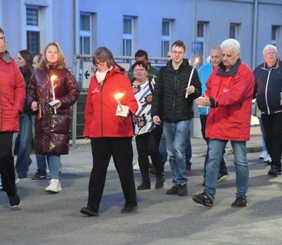 Menschen in Winterjacken halten brennende Kerzen in der Hand, während sie eine Straße entlang gehen. Sie sind zu einem Kerzenlicht-Gedenken versammelt.