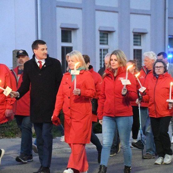 Eine Gruppe von Menschen in roten Mänteln hält brennende Kerzen, während sie am Abend gehen. Dahinter steht ein Gebäude mit vielen Fenstern.