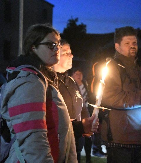 Drei Menschen halten brennende Kerzen und stehen bei Nacht im Freien. Die Frau vorn trägt eine Jacke mit roten Streifen und eine Brille. Der Mann neben ihr trägt eine Jacke mit einem orangefarbenen Streifen. Der Mann rechts trägt eine braune Jacke. Bäume und Gebäude sind im Hintergrund.