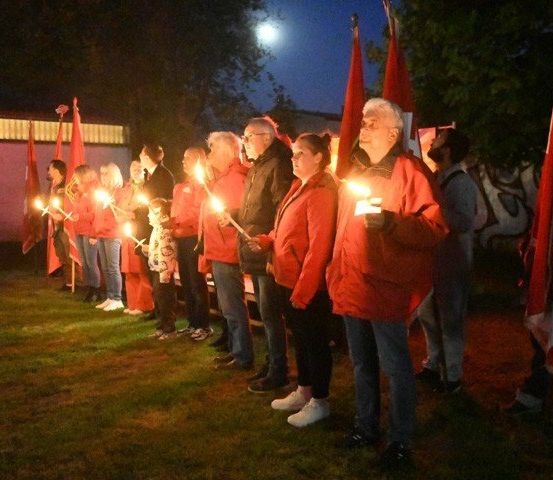Eine Gruppe von Menschen in roten Jacken hält brennende Kerzen in einer Reihe, unter einem Vollmond, mit Fahnen dahinter.
