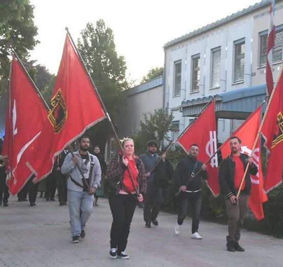 Eine Gruppe von Menschen läuft zusammen und trägt rote Flaggen mit Symbolen. Vor ihnen spielt eine Band. Der Weg ist von Gebäuden und Grünflächen gesäumt.