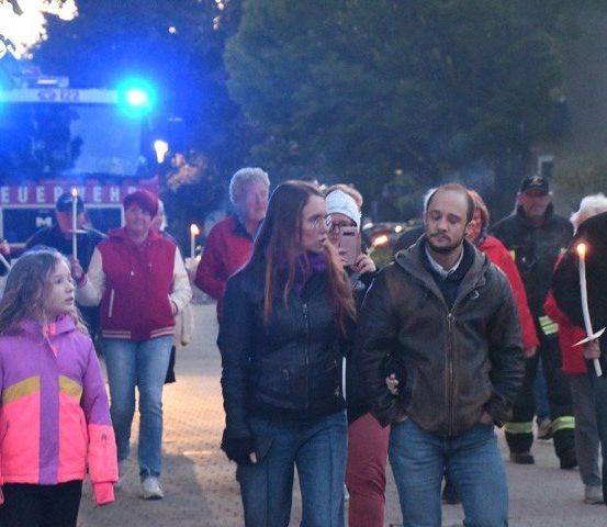 Eine Gruppe von Menschen, die Kerzen halten, marschiert bei Nacht auf der Straße, mit einem Feuerwehrauto im Hintergrund.