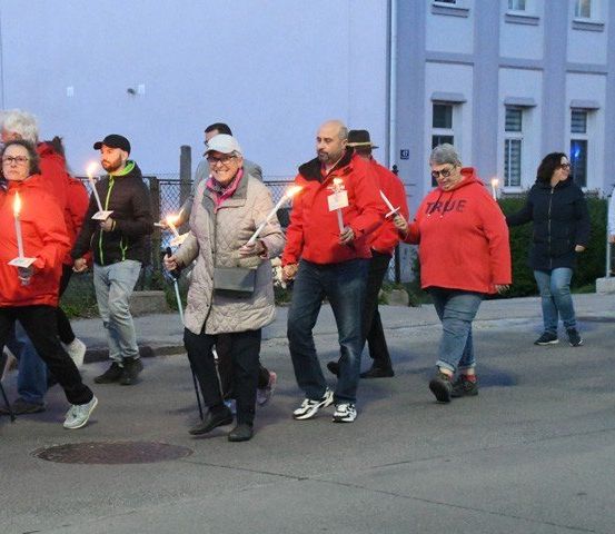 Eine Gruppe von Menschen mit brennenden Kerzen in Winterkleidung geht auf einer Straße, ein Mann in Feuerwehruniform.