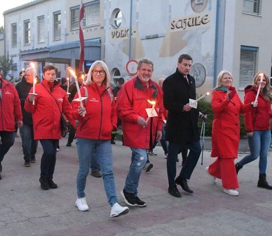 Eine Gruppe von Menschen in roten Jacken, die mit brennenden Kerzen in den Händen vor einem Gebäude gehen.