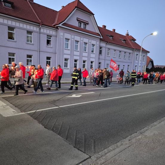 Eine Gruppe von Menschen in roten Jacken und Kerzen marschiert vor einem Gebäude mit einem roten Dach und Fenstern. Einige halten rote Banner. Einige von ihnen tragen Hüte. Das Gebäude hat eine Straßenlaterne vor sich.