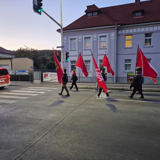 Eine Gruppe von Menschen geht mit roten Fahnen über eine Straße, mit einem Gebäude und einer Ampel im Hintergrund.