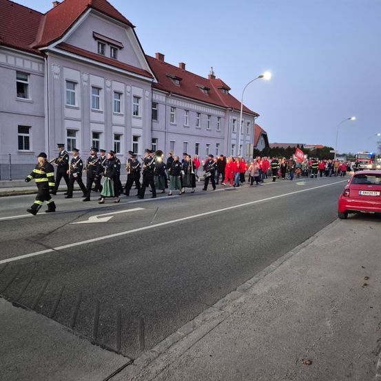 Eine Blaskapelle mit Instrumenten und eine Gruppe von Menschen in roten Kleidern gehen die Straße entlang. Ein rotes Auto ist am Straßenrand geparkt.