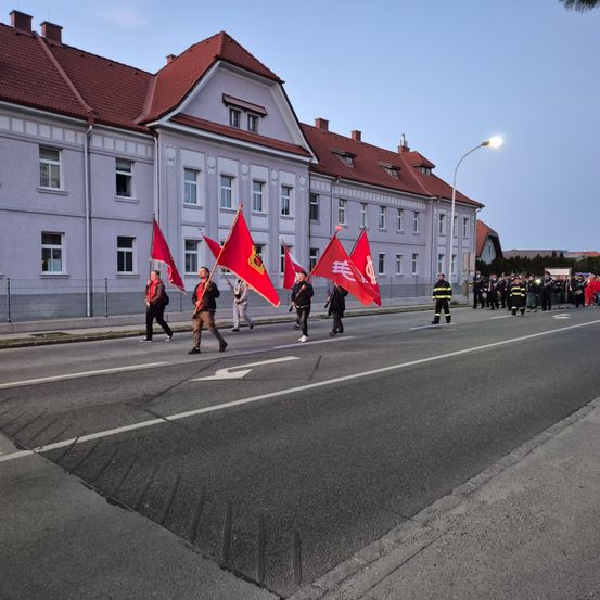 Eine Gruppe von Menschen geht mit roten Fahnen in der Hand die Straße entlang. Sie stehen vor einem großen Gebäude mit einem roten Dach. Andere stehen am Straßenrand.