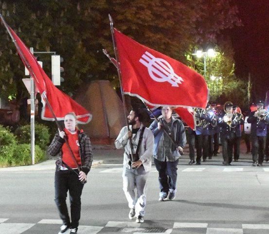 Eine Gruppe von Menschen, die Flaggen halten, geht nachts auf der Straße, während eine Band hinter ihnen spielt.