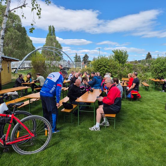 Eine Gruppe von Radfahrern und Menschen sitzt auf Bänken in einem Park. Einige schauen auf ihre Handys, während andere sprechen. Ein Fahrrad ist in der Nähe geparkt.
