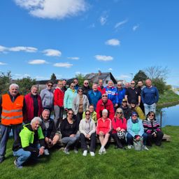 Eine Gruppe von Menschen posiert für ein Foto vor einem Kanal, in verschiedenen Jacken und Sonnenbrillen. Einige sitzen auf einer Holzbank, während andere hinter ihnen stehen. Hinter der Gruppe sind Häuser und Bäume zu sehen.