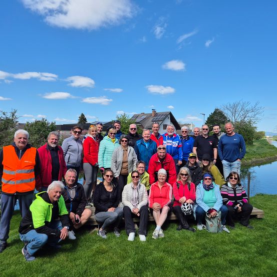 Eine Gruppe von Menschen posiert für ein Foto vor einem Kanal, in verschiedenen Jacken und Sonnenbrillen. Einige sitzen auf einer Holzbank, während andere hinter ihnen stehen. Hinter der Gruppe sind Häuser und Bäume zu sehen.