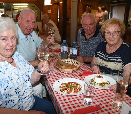 Vier ältere Personen sitzen an einem Tisch, lächeln und halten Weingläser in den Händen. Sie sind von Tellern mit Essen, Wasserflaschen und einem Korb mit Brot umgeben. Der Tisch ist mit einer rot-weiß karierten Tischdecke bedeckt.