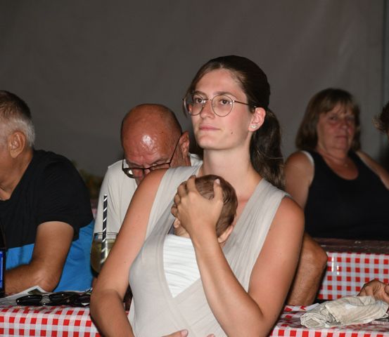 Eine Frau mit Brille hält ein Baby in einem Tragetuch, während sie mit anderen Leuten an einem Tisch sitzt.