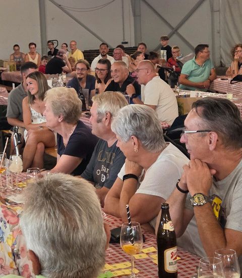 Eine Gruppe von Menschen sitzt an Tischen in einem großen Raum, einige unterhalten sich, und es gibt Flaschen und Gläser auf den Tischen.