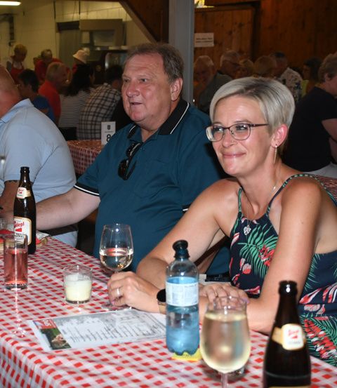 Ein Mann und eine Frau sitzen an einem Tisch mit Bier- und Weinflaschen und lächeln in die Kamera. Sie sind von anderen Leuten umgeben, die an Tischen in einem Restaurant sitzen.