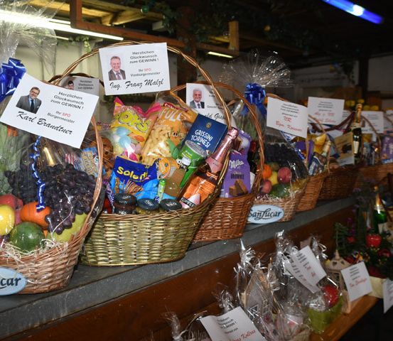 Eine Reihe von Körben, gefüllt mit verschiedenen Früchten, Snacks und Blumen, mit einem Schild, das 'SanLucar' auf dem linken Korb steht. Die Körbe sind in Plastik eingewickelt und auf einem Holzregal platziert.