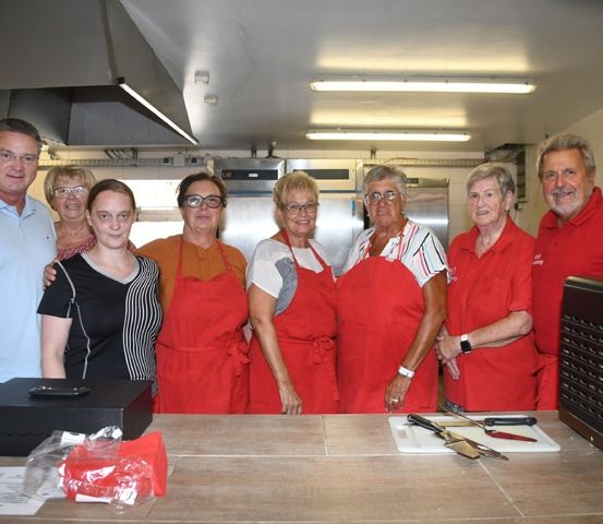 Eine Gruppe von Menschen in roten Schürzen steht in einer Küche, lächelt und posiert für ein Foto. Ein Holztisch mit einer schwarzen Box und verschiedenen Gegenständen ist im Vordergrund.