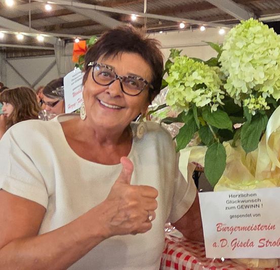 Eine Frau mit Brille und weißem Shirt hält einen Strauß grüner Blumen mit einem Schild, das Herzlichen Glückwunsch zum GEWINN 1 besagt. Sie gibt einen Daumen hoch.