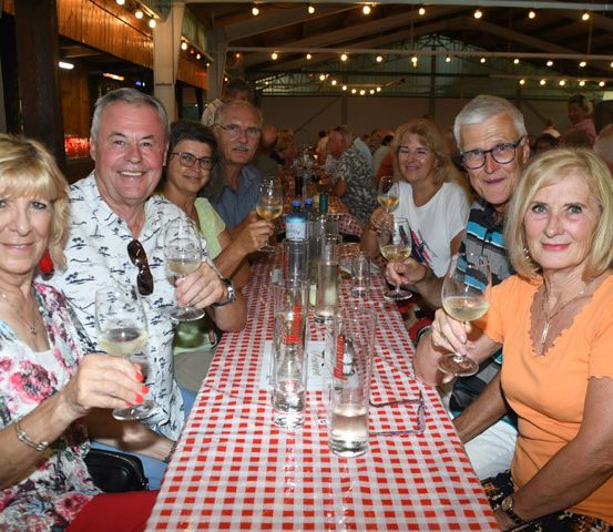 Eine Gruppe von Menschen sitzt an einem Tisch und lächelt, während sie Weingläser halten. Sie befinden sich wahrscheinlich bei einer Party oder einem gesellschaftlichen Ereignis.