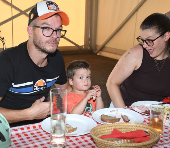 Eine Familie mit drei Personen, einschließlich eines kleinen Jungen, sitzt an einem Tisch mit rot-weiß kariertem Tischtuch und genießt eine Mahlzeit. Der Mann trägt eine Baseballkappe, und die Frau trägt eine Brille. Ein Fahrrad ist hinter ihnen geparkt.