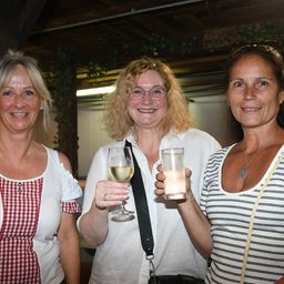 Drei Frauen stehen zusammen, zwei halten Getränke in der Hand und lächeln in einer festlichen Umgebung. Die Frau auf der linken Seite trägt ein rot-weiß kariertes Oberteil. Die Frau in der Mitte hält ein Weinglas und die Frau auf der rechten Seite ein Glas mit einem Getränk.