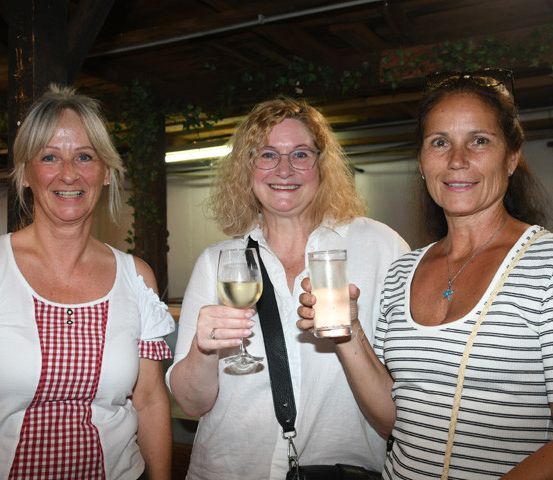 Drei Frauen stehen zusammen, zwei halten Getränke in der Hand und lächeln in einer festlichen Umgebung. Die Frau auf der linken Seite trägt ein rot-weiß kariertes Oberteil. Die Frau in der Mitte hält ein Weinglas und die Frau auf der rechten Seite ein Glas mit einem Getränk.