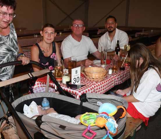 Eine Familie sitzt an einem Tisch in einem Restaurant, mit einem Baby in einem Kinderwagen im Vordergrund. Der Tisch hat eine rot-weiß karierte Tischdecke, und verschiedene Getränke und Essensartikel sind vorhanden.