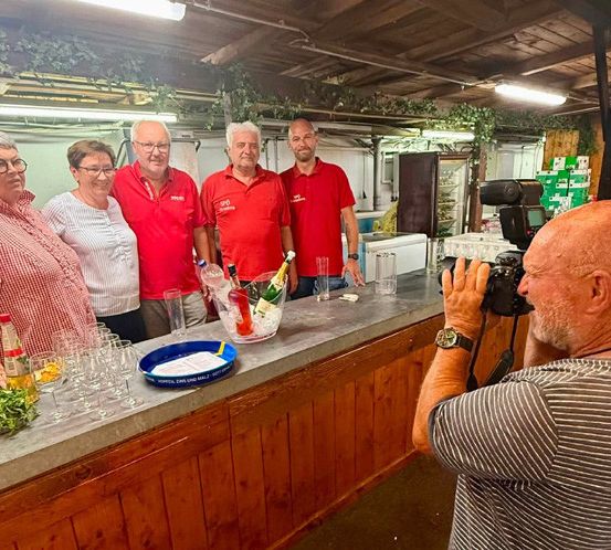 Eine Gruppe von Menschen in roten Shirts steht hinter einer Bartheke, während ein Mann mit einer Kamera ein Foto macht. Die Theke hat Flaschen, Gläser und eine Schüssel.