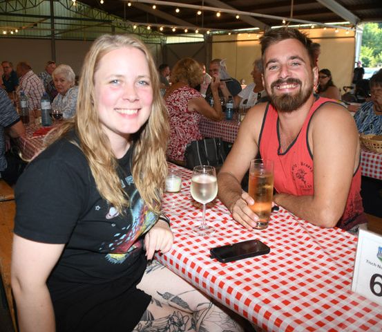 Ein Mann und eine Frau sitzen an einem Tisch in einem Restaurant, lächeln und posieren für ein Foto. Sie haben Getränke vor sich, und der Tisch hat die Nummer 6. Hinter ihnen sitzen andere Leute an Tischen, und es gibt Flaschen und Gläser auf den Tischen.