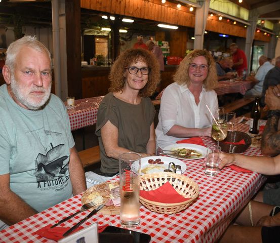 Vier Menschen sitzen an einem Tisch in einem Restaurant, lächeln und genießen ihr Essen. Ein Mann mit Bart und eine Frau mit Brille gehören dazu. Der Tisch ist mit Tellern, Gläsern und einem Korb gedeckt. Im Hintergrund sind andere Tische und sitzende Menschen zu sehen.