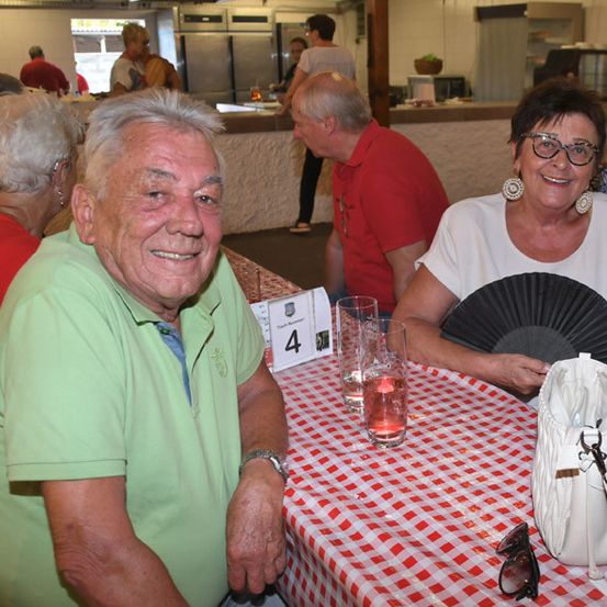 Ein älterer Mann und eine ältere Frau sitzen an einem Tisch mit rot-weiß kariertem Tischtuch und lächeln. Der Mann trägt ein grünes Poloshirt und die Frau eine weiße Bluse und eine Brille. Eine Handtasche und ein Fächer liegen auf dem Tisch. Hinter ihnen stehen und sitzen Menschen in einem Restaurant.