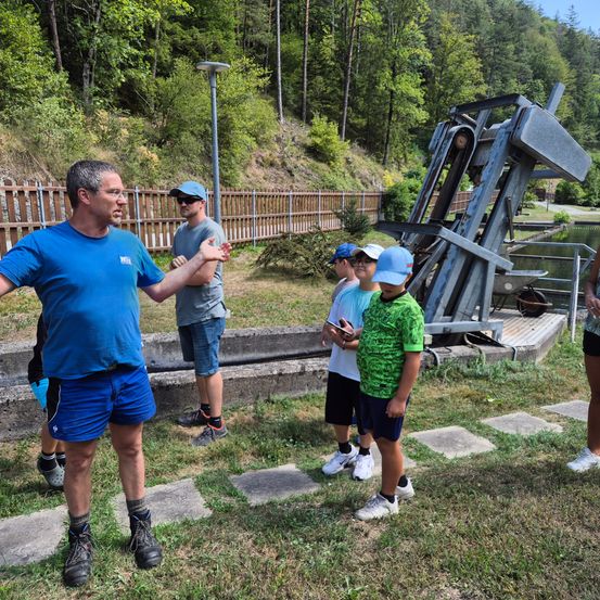 Eine Gruppe von Menschen, darunter ein Mann in einem blauen T-Shirt, steht in der Nähe einer großen mechanischen Struktur auf einem Grasfeld mit einem Holzzaun und Bäumen im Hintergrund.
