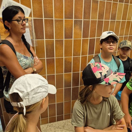 Eine Frau mit Brille und Rucksack steht neben einer gefliesten Wand und schaut eine Gruppe von Kindern an, die Mützen und Rucksäcke tragen. Die Kinder schauen in verschiedene Richtungen.