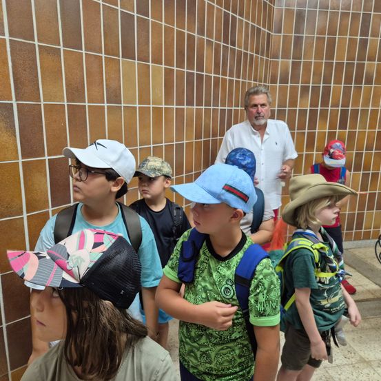 Eine Gruppe von Kindern in Hüten und Rucksäcken steht in einem gefliesten Flur, mit einem Mann in einem weißen Hemd und einer Frau in einer roten Mütze im Hintergrund.
