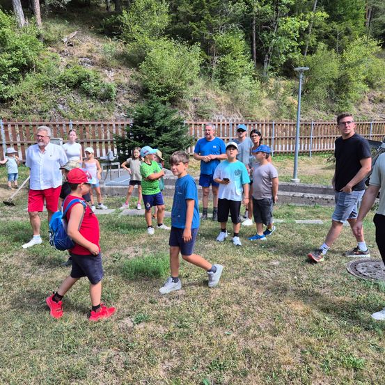 Eine Gruppe von Menschen, einschließlich Erwachsener und Kindern, versammelt sich auf einem Rasen mit einem Zaun und Bäumen im Hintergrund. Einige tragen Hüte und Turnschuhe.