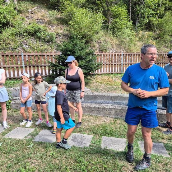Eine Gruppe von Menschen, darunter Kinder, steht auf einem Rasen. Ein erwachsener Mann in einem blauen T-Shirt spricht zu ihnen. Hinter ihnen befindet sich ein Holzzaun und ein Hang mit Bäumen.