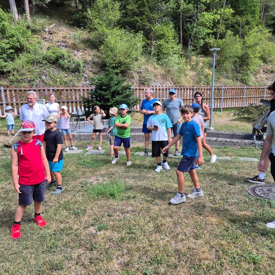 Eine Gruppe von Menschen, einschließlich Kindern und Erwachsenen, steht in einem Grasbereich, möglicherweise in einem Park, mit einem Holzzaun und Bäumen im Hintergrund. Einige tragen Hüte und Turnschuhe.