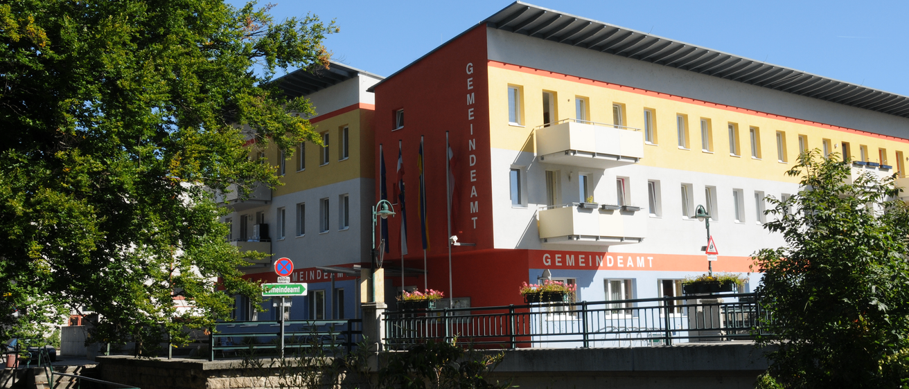 Ein Gebäude mit roten, gelben und weißen Wänden, es hat mehrere Flaggen, Balkone und Topfpflanzen. Ein Straßenschild und eine Laterne sind davor.