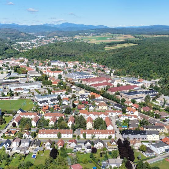 Luftaufnahme einer Stadt mit vielen Gebäuden, umgeben von grünen Bäumen und Bergen in der Ferne. Die Stadt liegt in einem Tal.
