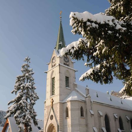 Eine schneebedeckte Kirche mit hohem Turm, einer Uhr und einem Kreuz oben steht vor blauem Himmel mit Bäumen darum herum.