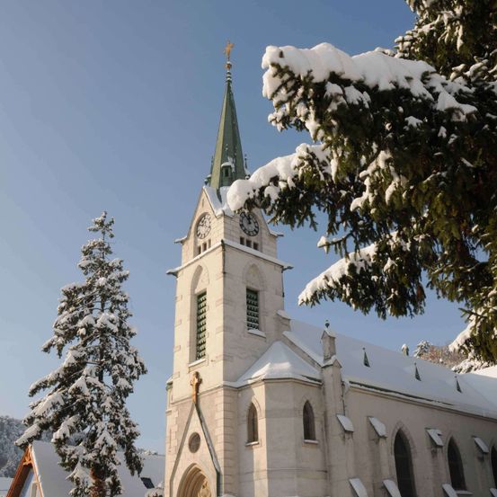 Eine schneebedeckte Kirche mit hohem Turm, einer Uhr und einem Kreuz oben steht vor blauem Himmel mit Bäumen darum herum.