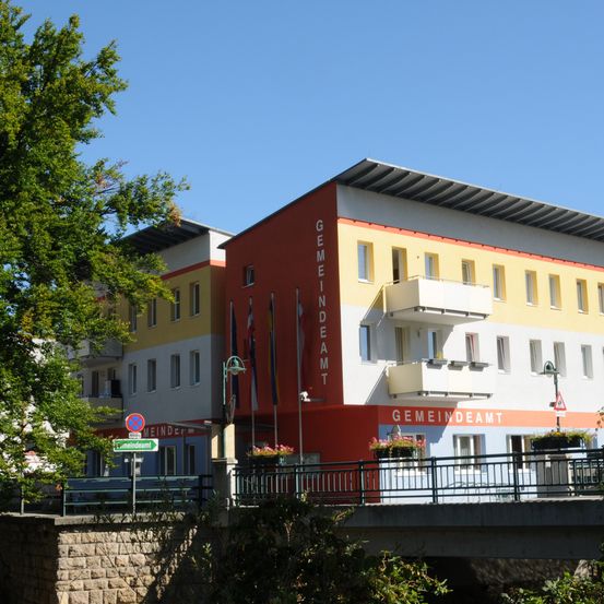 Ein mehrfarbiges Gebäude mit mehreren Flaggen, beschriftet mit 'Gemeindeamt', steht neben einer Brücke mit Bäumen und einem Schild.