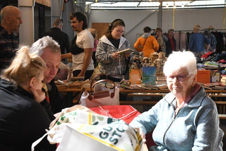 People are shopping in an indoor market with stalls selling books and other items. A woman reads a book while others converse.