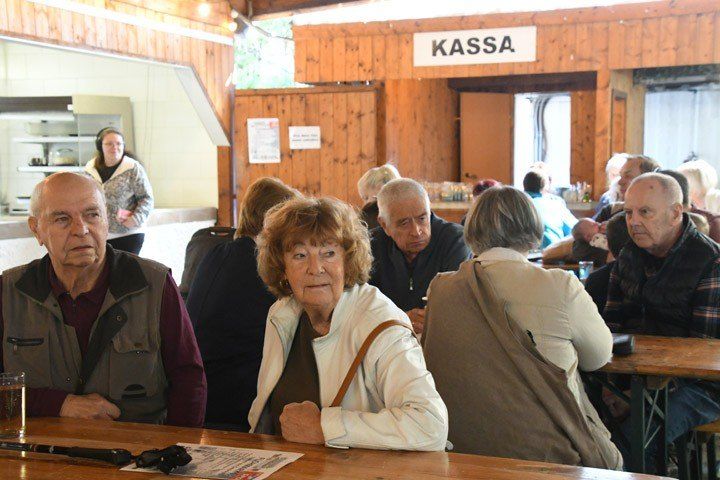 A group of older people sit around a wooden table in a restaurant with a sign that says KASS. A woman with headphones stands near a counter.