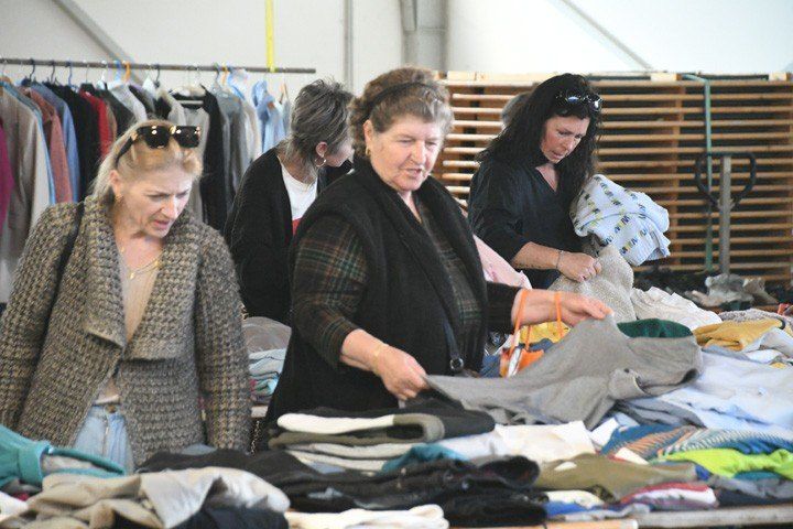 Four women, some wearing sunglasses, are sorting through clothes on a table in a clothing store.