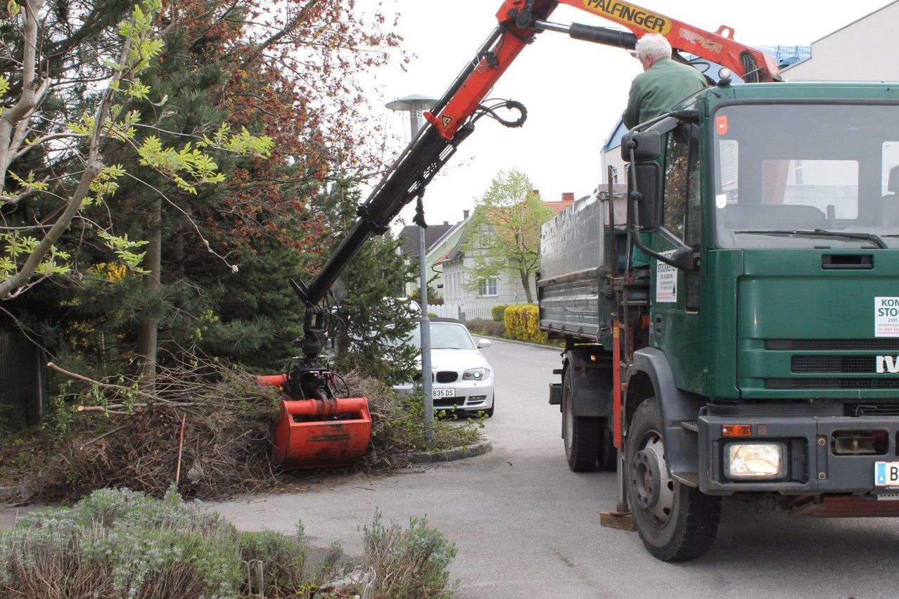Ein großer roter und schwarzer Kran steht neben einem grünen Lastwagen. Ein Mann sitzt im Lastwagen und beobachtet, wie der Kran Äste vom Boden hebt. In der Nähe ist ein weißer Wagen geparkt, und ein Haus ist im Hintergrund zu sehen.